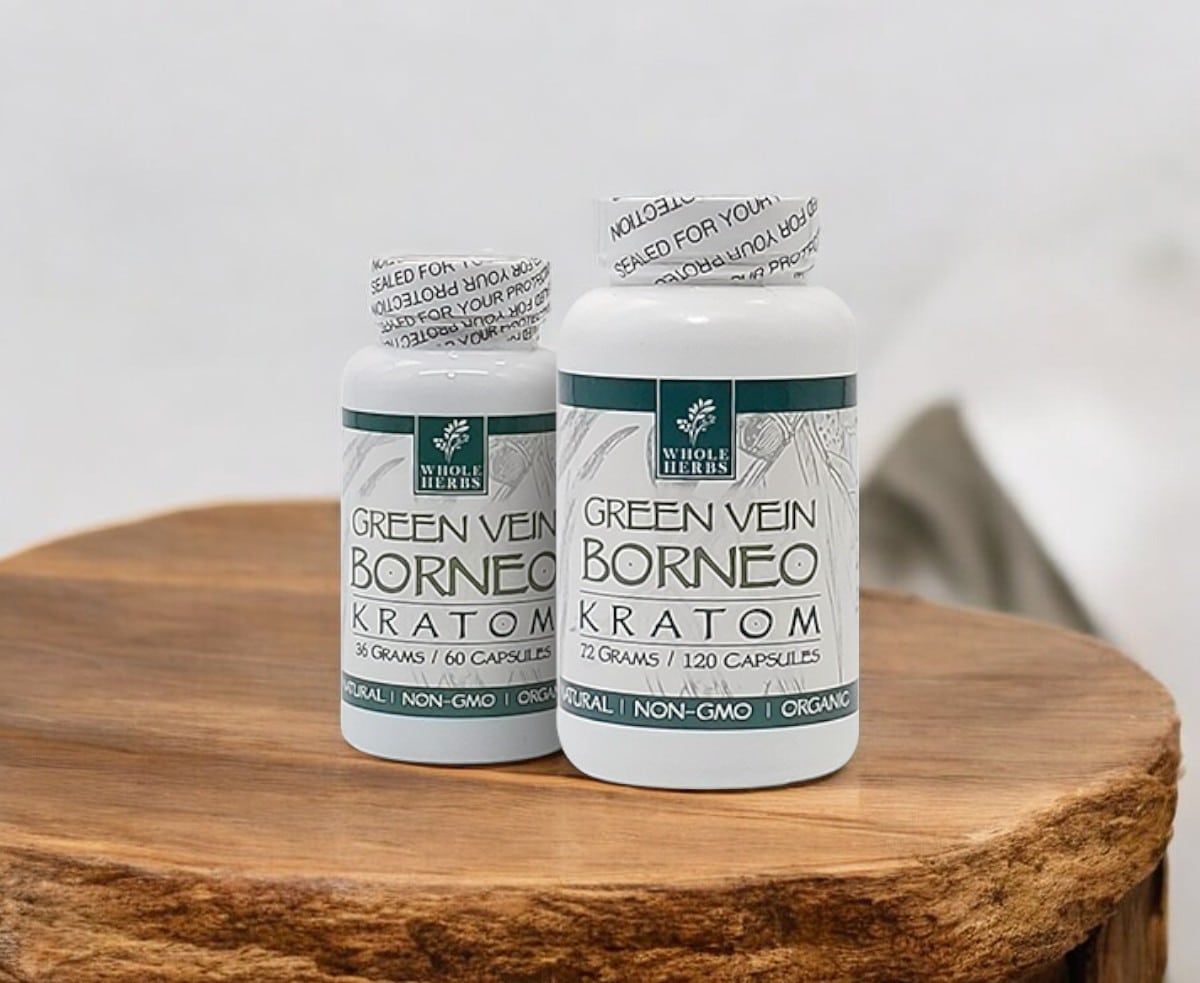 Displayed on a wooden surface are two bottles of Whole Herbs – Green Vein Borneo Capsules. Both carry labels indicating they are organic and non-GMO, with contents of 36 grams in one bottle and 72 grams in the other.