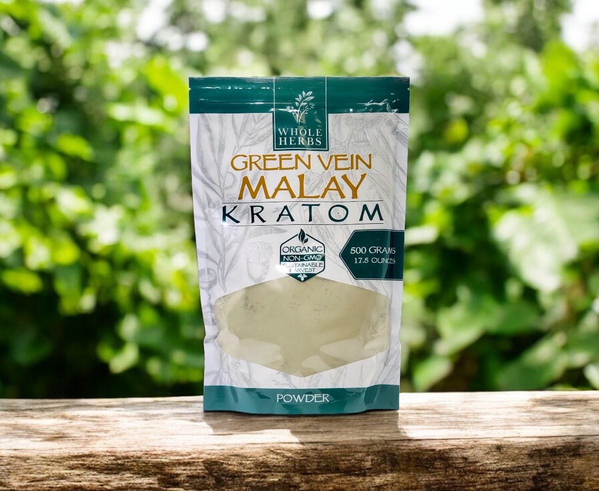 A bag of Whole Herbs – Green Vein Malay Kratom Powder sits on a wooden surface, its 500 grams distinctly highlighted against a softly blurred green backdrop.