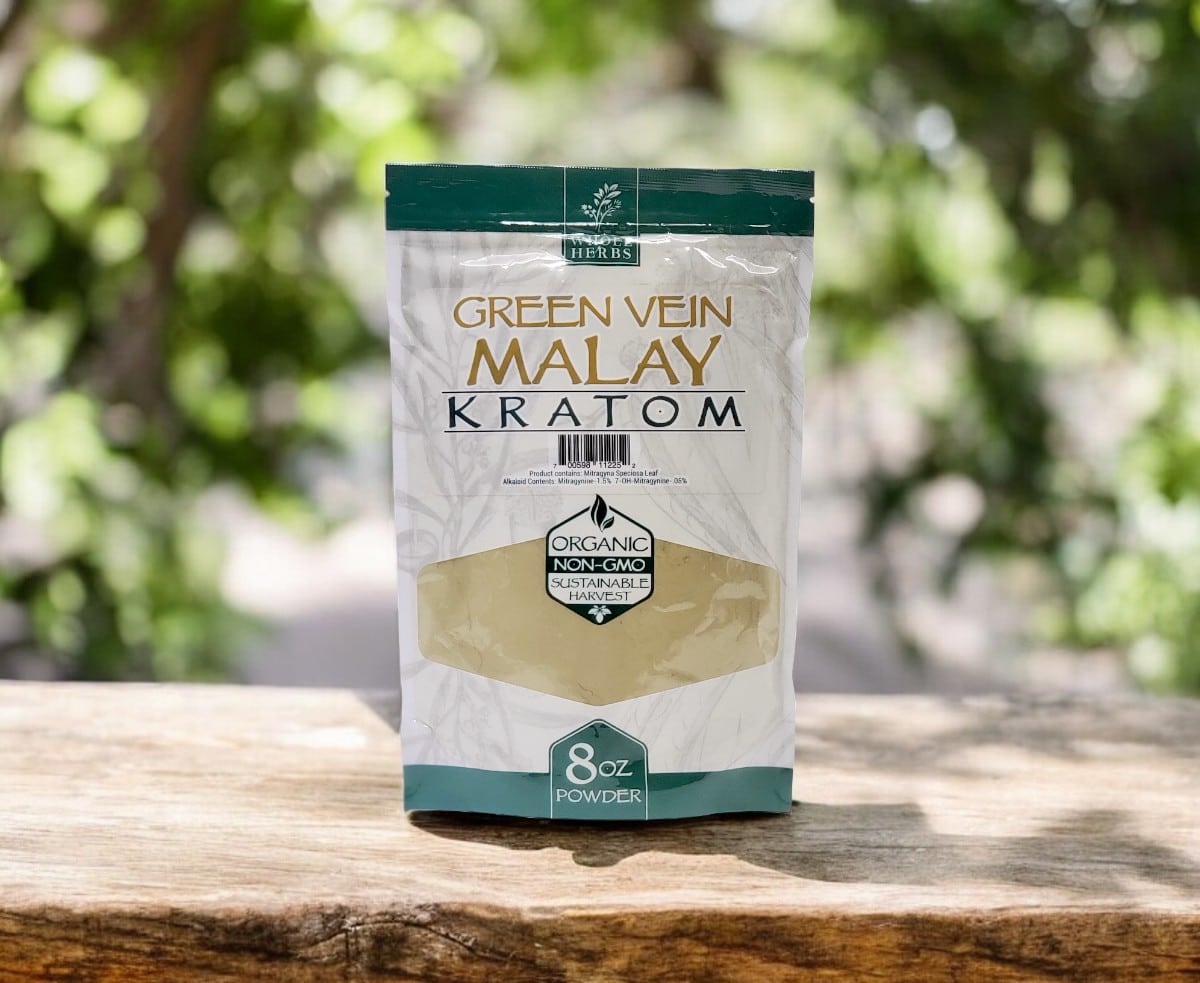 Front view of an 8 oz green and white package of Whole Herbs – Green Vein Malay Kratom Powder, labeled as non-GMO, resting on a wooden surface with trees in the background.
