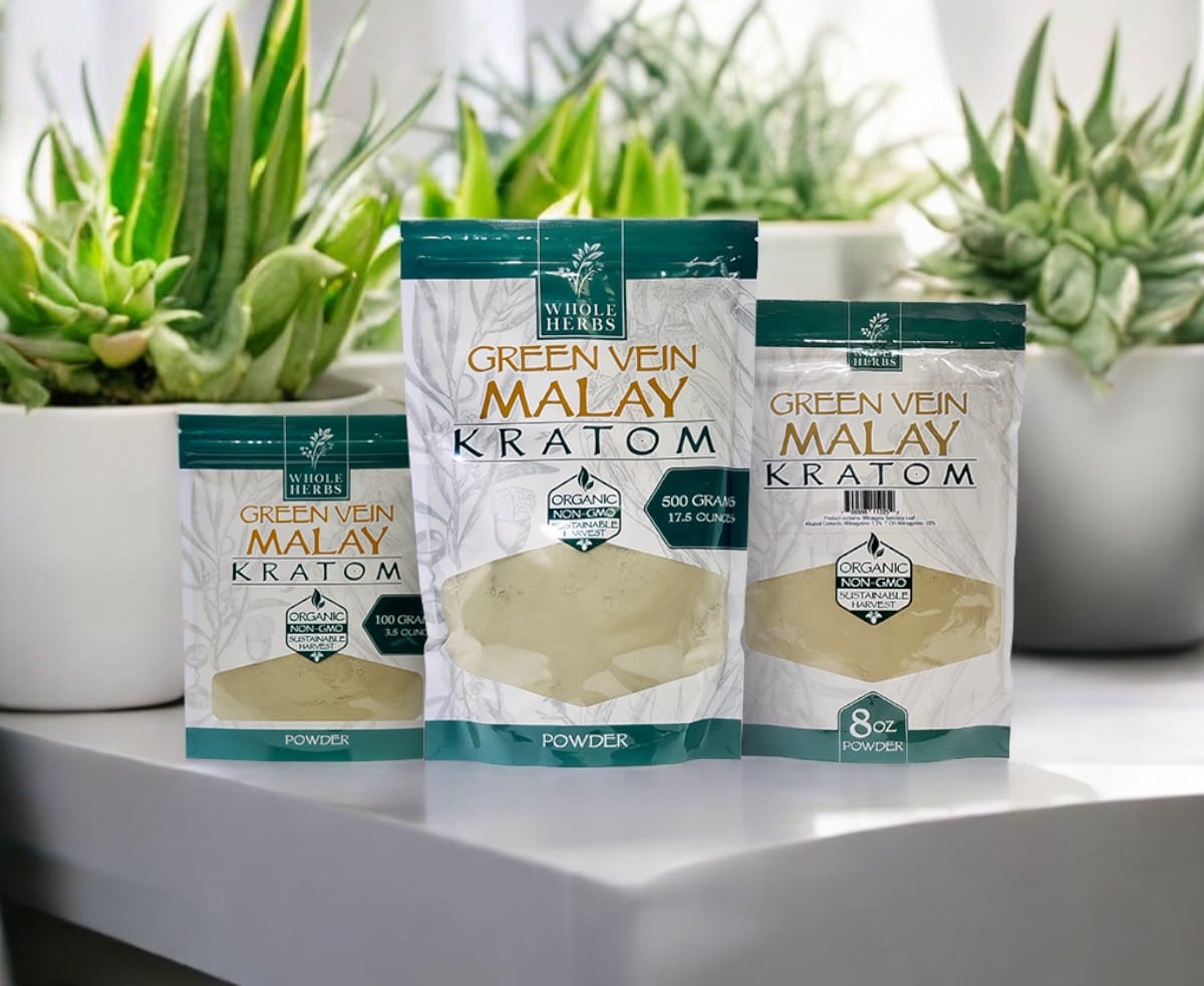 Three packs of Whole Herbs – Green Vein Malay Kratom Powder are arranged on a surface, with potted succulent plants providing a natural backdrop.