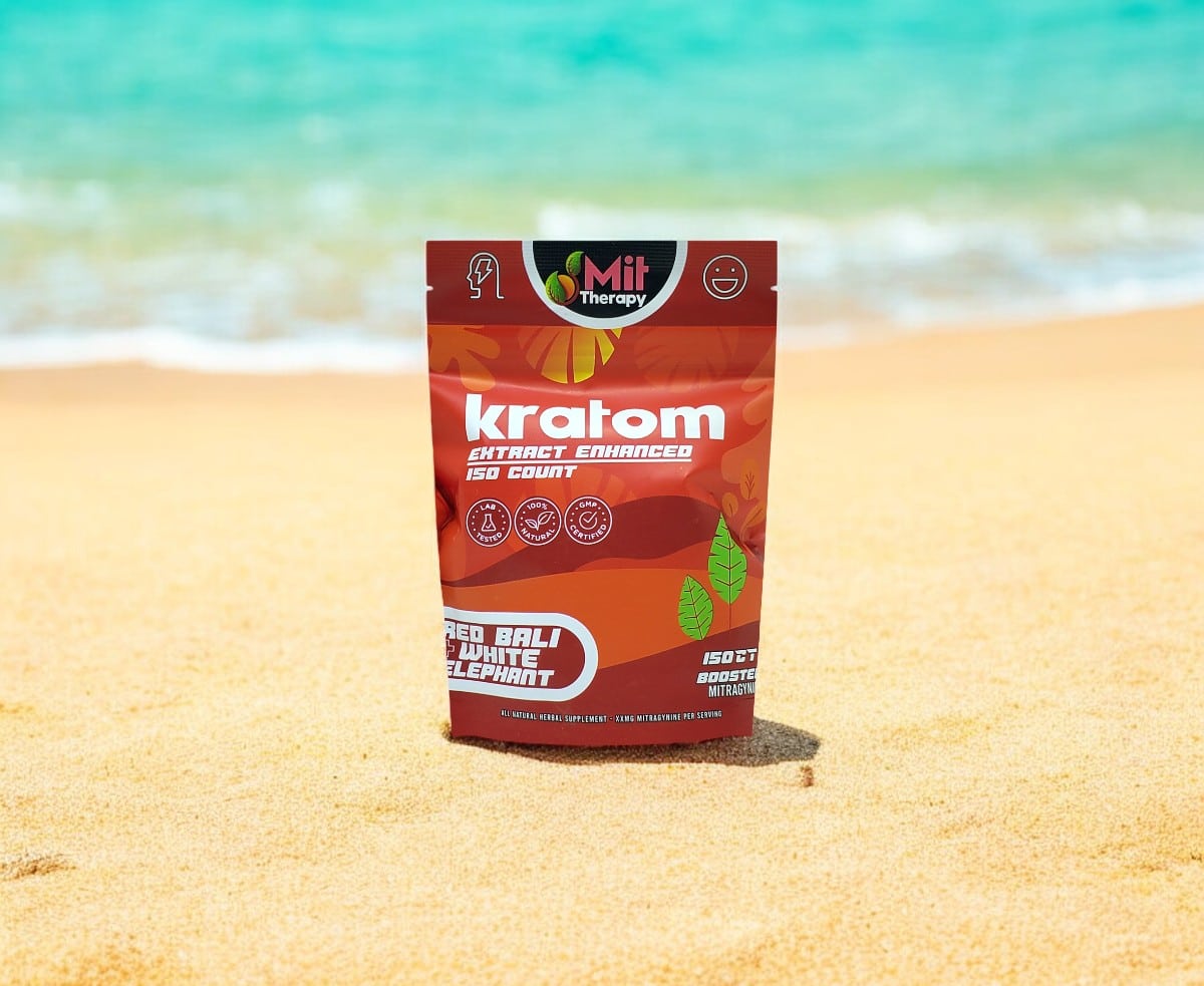 A package of Mit Therapy Red Bali + White Elephant Capsules stands upright on a sandy beach with the ocean in the background, inviting relaxation and tranquility reminiscent of a serene therapy session.