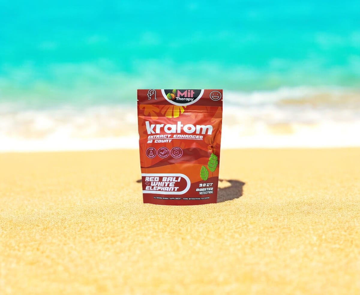 A package of Mit Therapy Red Bali + White Elephant Capsules stands upright on a sandy beach with a clear blue sea in the background, offering a serene escape akin to a tranquil therapy for the mind and soul.