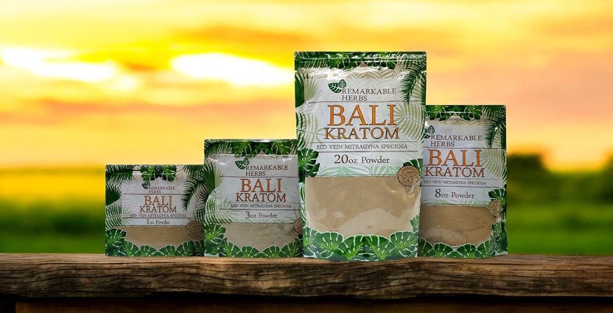 Four bags of Remarkable Herbs Red Bali Kratom Powder are elegantly displayed on a wooden surface, with a blurred outdoor background adding to their allure.