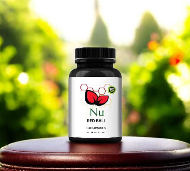 A bottle of Nu – Red Bali capsules sits outside on a wooden surface, contrasting with the vibrant greenery in the blurred background.